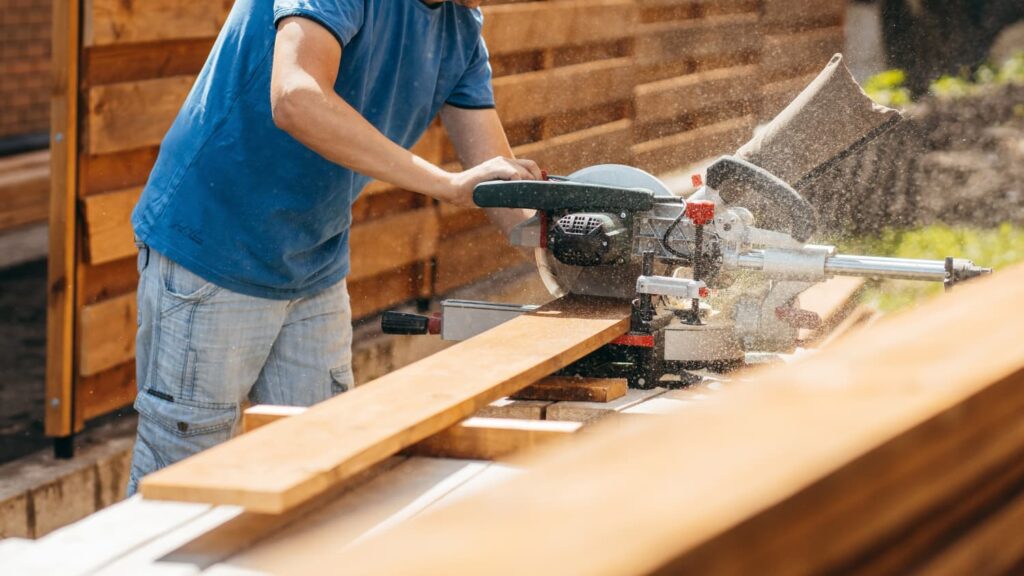A man uses a miter saw to cut lumber outdoors during a deck or fence building project.