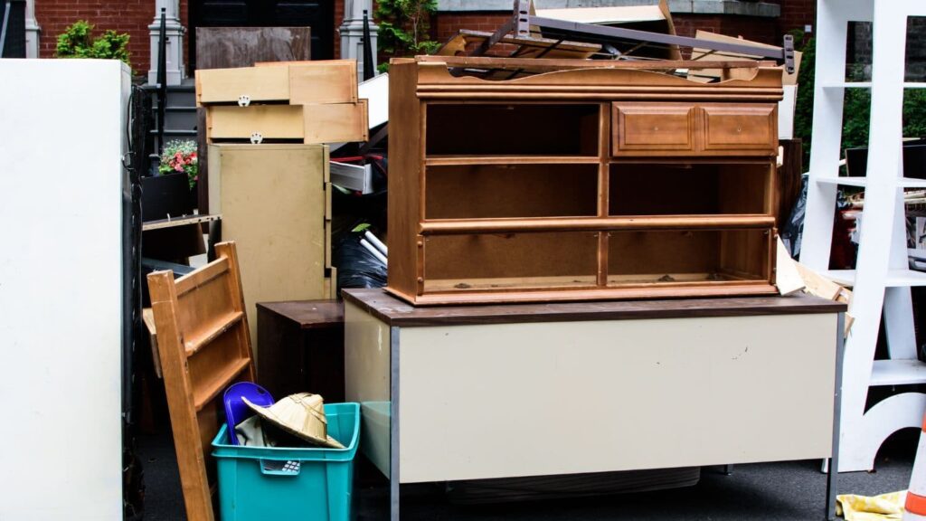 A pile of discarded furniture and household items on a curb, including a wooden dresser, a filing cabinet, a chest of drawers, a wooden ladder-back chair, plastic storage bins, and a straw hat, ready for a moving sale or trash pickup.
